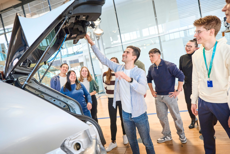 Visitor stands at the vehicle and tests functions on the vehicle. Group of visitors with guide stands in the background.