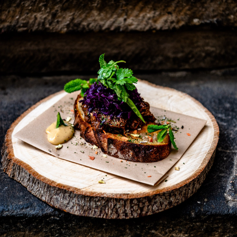 Auf einer Baumscheibe ist eine Scheibe Krustenbrot mit einer Variation aus Fleisch und Salat trapiert.
