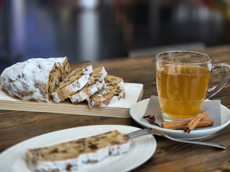 A slice of stollen on a plate. Next to it is a cup of mulled wine, the saucer decorated with a napkin, cinnamon and cloves